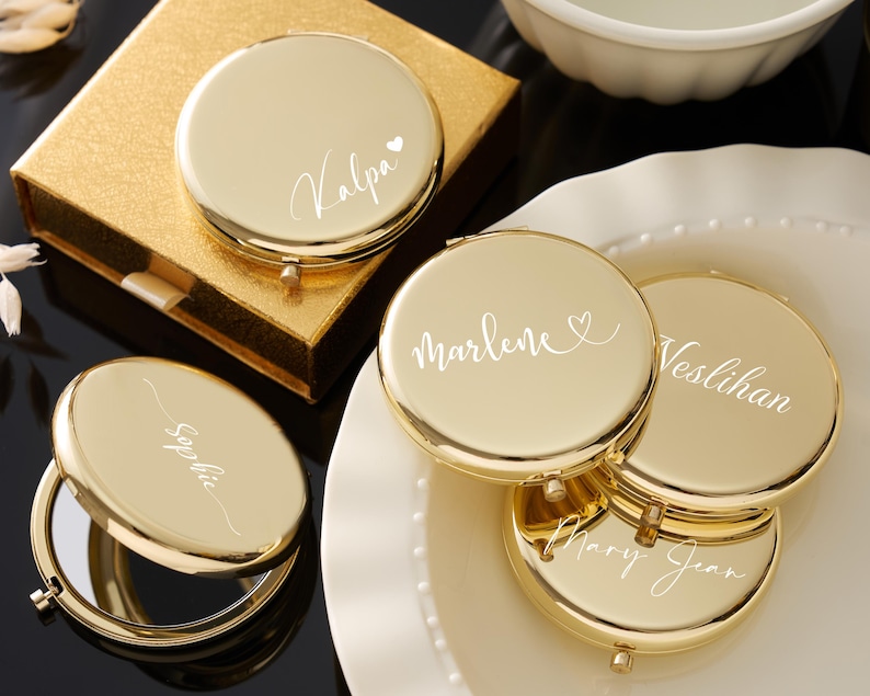 May include: Gold-colored compact mirrors with personalized names in white script. One mirror is open, revealing a reflective surface. The mirrors are on a white plate and a gold gift box. The names include "Kalpa", "Sophie", "Marlene", "Veslihan", and "Mary Jean".