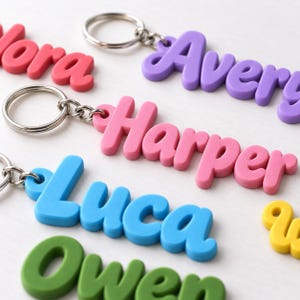 May include: A collection of personalized keychains featuring raised, rounded letters in various colors including purple, pink, blue, green, and yellow. Each keychain is attached to a silver-toned metal split ring and displays a name.