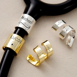 Pu&ograve; includere: Uno stetoscopio nero con anelli in metallo personalizzati. Gli anelli sono argentati e dorati, incisi con nomi e simboli. Un anello dice "Michael" con un cuore, un altro dice "Lucas" con un'icona di flacone di pillole. Altri anelli dicono "Buster" e "Victoria".