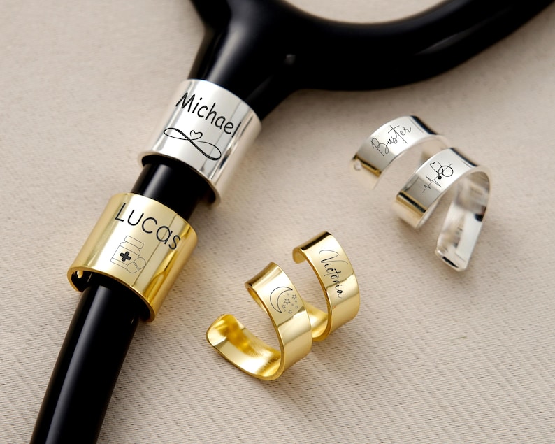 Pu&ograve; includere: Uno stetoscopio nero con anelli in metallo personalizzati. Gli anelli sono argentati e dorati, incisi con nomi e simboli. Un anello dice "Michael" con un cuore, un altro dice "Lucas" con un'icona di flacone di pillole. Altri anelli dicono "Buster" e "Victoria".