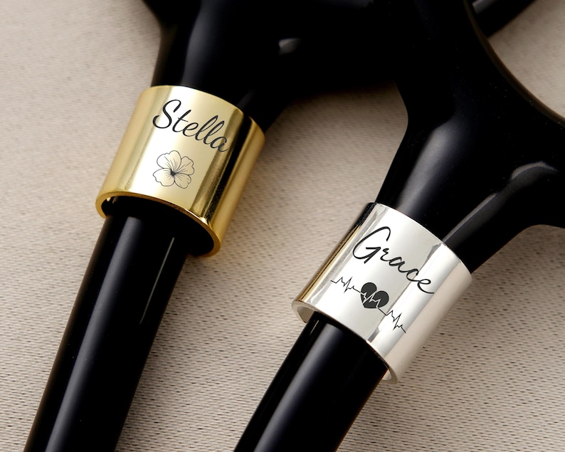 Pu&ograve; includere: Primo piano di due stetoscopi con anelli personalizzati. Un anello &egrave; dorato con il nome "Stella" e un fiore, l'altro &egrave; argentato con il nome "Grace", un cuore e una linea del battito cardiaco.