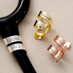 Pu&ograve; includere: Uno stetoscopio nero con ciondoli personalizzati per stetoscopio in metallo oro, argento e oro rosa. I ciondoli sono incisi con nomi, un sole, un battito cardiaco, un simbolo di stetoscopio e un motivo floreale.