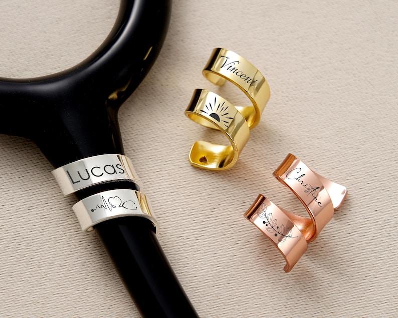 Pu&ograve; includere: Uno stetoscopio nero con ciondoli personalizzati per stetoscopio in metallo oro, argento e oro rosa. I ciondoli sono incisi con nomi, un sole, un battito cardiaco, un simbolo di stetoscopio e un motivo floreale.