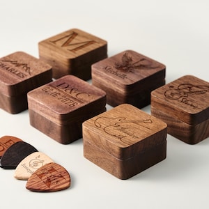 Puede incluir: Una colección de cajas de púas de guitarra de madera personalizadas y púas de guitarra. Las cajas cuadradas tienen diseños y nombres grabados. Las púas son de varios colores y diseños. Las cajas son de madera oscura.