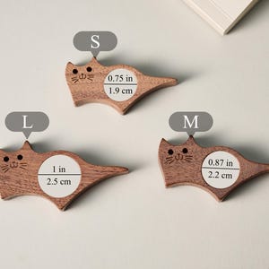 May include: Three wooden cat-shaped items in varying sizes. The smallest is 0.75 inches (1.9 cm) wide, the medium is 0.87 inches (2.2 cm) wide, and the largest is 1 inch (2.5 cm) wide. Each has a carved face.