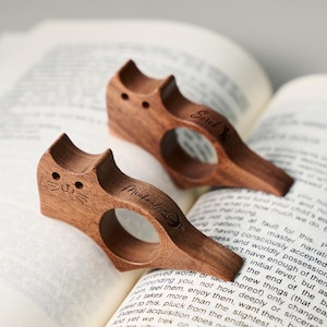 Peut inclure: Deux serre-livres en bois en forme de chat, posés sur un livre ouvert. Les supports sont de couleur marron chaud, avec des visages de chat gravés et des noms. Les pages du livre sont remplies de texte.