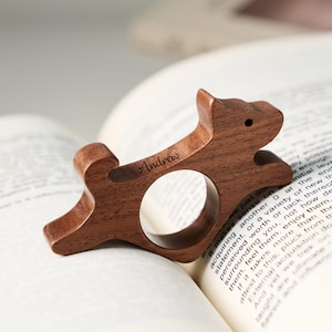 May include: A wooden dog-shaped bookmark with the name "Andrew" engraved on it. The bookmark is made of a dark brown wood and has a circular opening. It is resting on the pages of an open book.