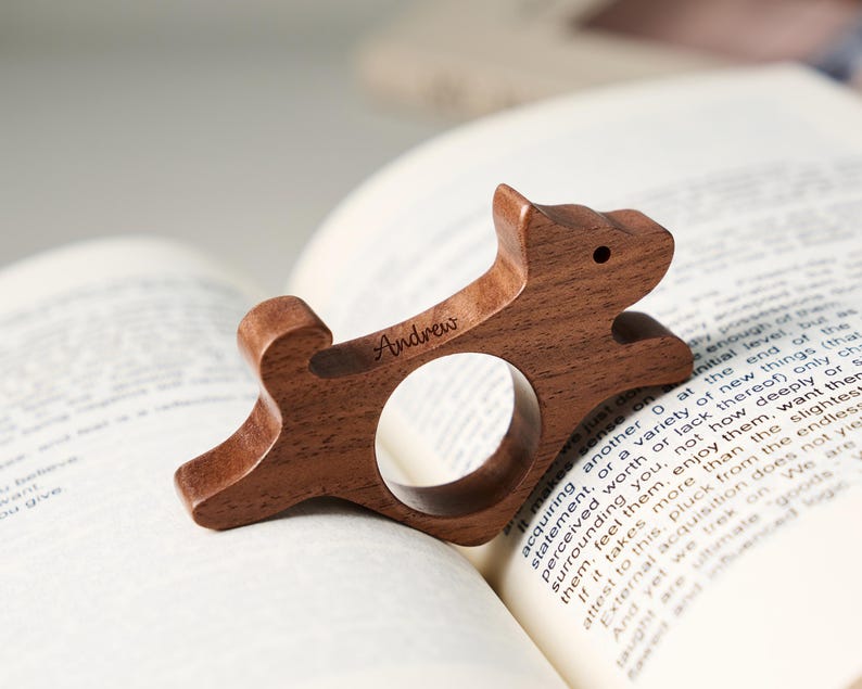 May include: A wooden dog-shaped bookmark with the name "Andrew" engraved on it. The bookmark is made of a dark brown wood and has a circular opening. It is resting on the pages of an open book.