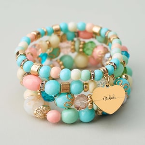 May include: A collection of pastel-coloured beaded bracelets, featuring pink, turquoise, and cream beads. Gold-toned accents and a heart charm with the name "Nicole" are visible. The bracelets use various bead sizes and shapes.