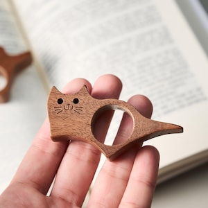 May include: A wooden cat-shaped book page holder. The brown holder has a circular opening for the finger and a tail-like extension. The cat's face is etched with eyes, whiskers, and a smiling mouth. Another holder is visible in the background.
