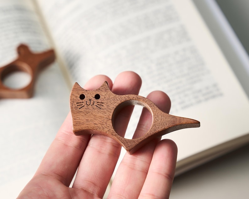 May include: A wooden cat-shaped book page holder. The brown holder has a circular opening for the finger and a tail-like extension. The cat's face is etched with eyes, whiskers, and a smiling mouth. Another holder is visible in the background.