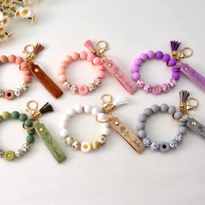 Könnte beinhalten: Eine Sammlung von Armband-Schlüsselanhängern in verschiedenen Farben, darunter Rosa, Grün, Lila und Grau. Jeder Schlüsselanhänger hat eine dekorative Blume und ein Lederetikett mit einem Namen. Die Schlüsselanhänger haben goldfarbene Hardware.