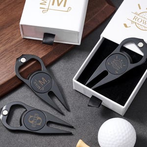 Könnte beinhalten: Drei mattschwarze Golf-Pitchgabeln aus Metall mit integrierten Flaschenöffnern. Jedes Werkzeug hat eine kreisförmige Mitte mit eingravierten Namen wie Miko, Steven und Rowan. Zwei weiße Schiebe-Geschenkboxen mit goldenem Text und ein weißer Golfball sind enthalten.