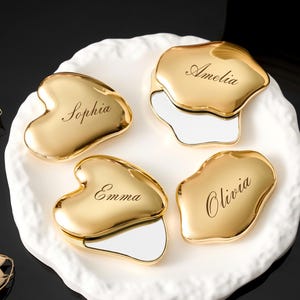 May include: Four gold-coloured compact mirrors, each with a different name: Sophia, Amelia, Emma, and Olivia. The mirrors are uniquely shaped and sit on a white, textured plate. The background is black.