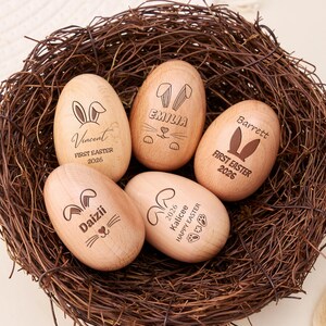 Könnte beinhalten: Fünf hölzerne Ostereier mit eingravierten Namen und Hasenmotiven in einem geflochtenen Nest. Die Eier tragen den Text "First Easter 2026" oder "Happy Easter". Die Eier sind hellbraun mit dunkelbraunen Gravuren.