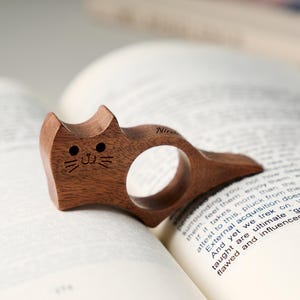 May include: A wooden cat-shaped book holder, resting on the pages of a book. The holder is carved from dark brown wood, featuring a circular finger hole and a smiling cat face with etched details. The word "Nir" is etched on the side.