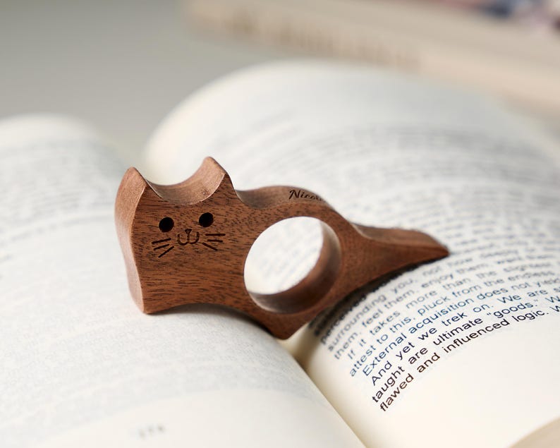 May include: A wooden cat-shaped book holder, resting on the pages of a book. The holder is carved from dark brown wood, featuring a circular finger hole and a smiling cat face with etched details. The word "Nir" is etched on the side.