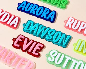 May include: A collection of colorful name tags in various colors, including blue, red, and green. Each tag features a different name in a bold, white font. The tags have a glossy finish and a small hole for attachment.