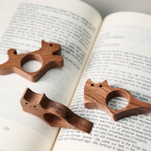 May include: Three wooden cat-shaped book page holders. One has the name "Megan" and a paw print engraved. The holders are made of dark wood and are resting on the pages of an open book.