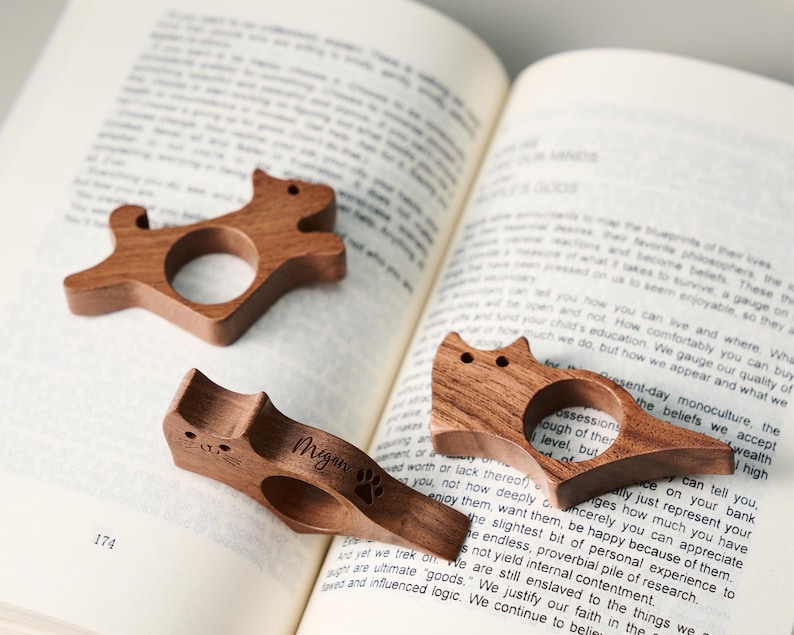 May include: Three wooden cat-shaped book page holders. One has the name "Megan" and a paw print engraved. The holders are made of dark wood and are resting on the pages of an open book.