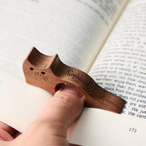 May include: A wooden cat-shaped book page holder, held open on a book. The holder is brown with the name "Sophia" and a heart design engraved on it. The book is open to pages 174 and 175.