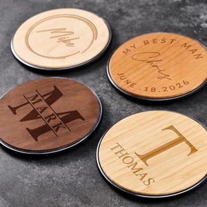 May include: Four personalised wooden coasters with silver rims. One coaster has "Mike" engraved, another says "My Best Man Chris June 18.2026", a third has "MARK" with a large initial, and the last says "THOMAS" with a large initial.