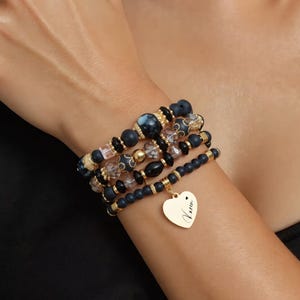 Peut inclure: Plusieurs bracelets de perles noirs, dorés et transparents. Les bracelets présentent différentes tailles et textures de perles, dont un charm en forme de cœur doré gravé du nom "Karen". Les bracelets sont portés sur un poignet.