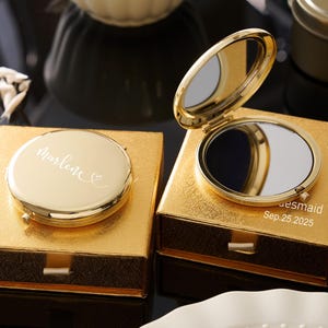 May include: Two gold compact mirrors, one open and one closed, sit on gold boxes. The closed mirror has the name "Marlene" in script. The open mirror reveals a reflective surface. The box of the open mirror has the text "bridesmaid Sep. 25.2025".