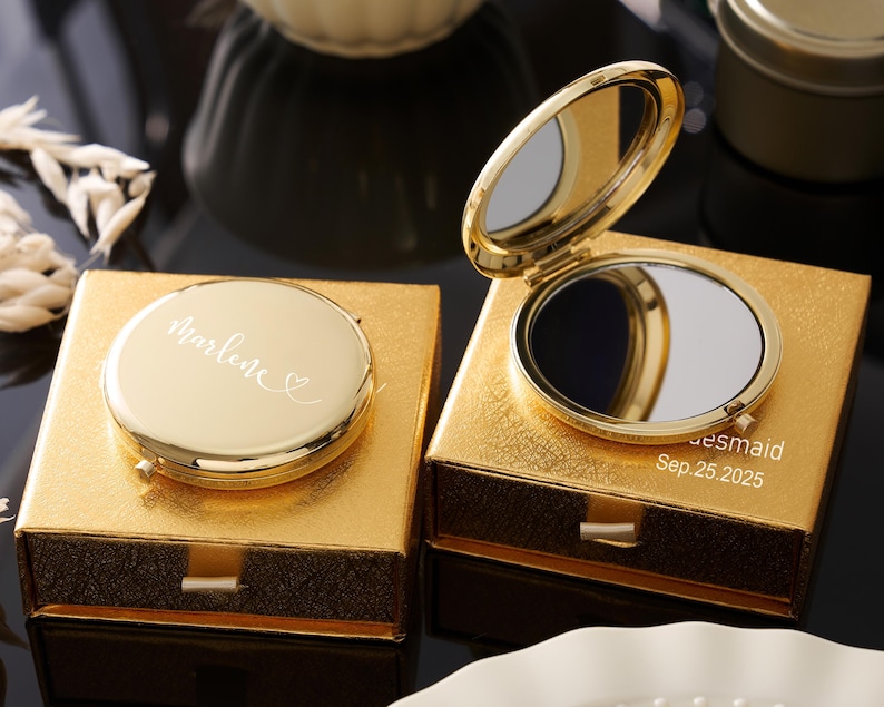 May include: Two gold compact mirrors, one open and one closed, sit on gold boxes. The closed mirror has the name "Marlene" in script. The open mirror reveals a reflective surface. The box of the open mirror has the text "bridesmaid Sep. 25.2025".