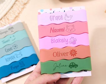 Könnte beinhalten: Ein Set aus fünf gewellten Silikonbändern in Helllila, Pink, Hellrosa, Orange und Grün, die jeweils mit einem Namen und einem kleinen Symbol wie einem Wal, Fuchs, einer Biene, einer Blume oder einem Auto versehen sind.