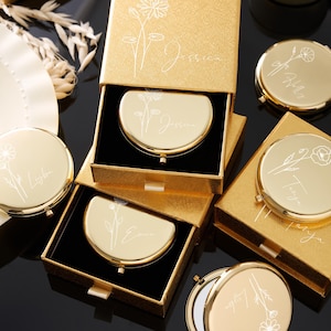 May include: Gold-colored compact mirrors with floral designs and personalized names are displayed in open boxes. The mirrors are round and have a hinged opening. The boxes are gold with a textured finish. The names "Jessica", "Emma", "Tanya", and "Lyla" are engraved on the mirrors.