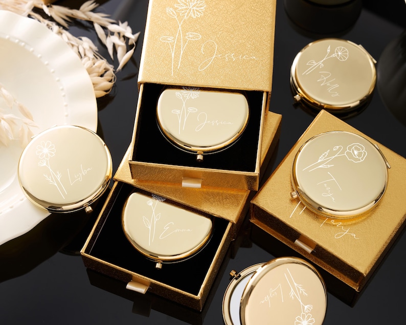 May include: Gold-colored compact mirrors with floral designs and personalized names are displayed in open boxes. The mirrors are round and have a hinged opening. The boxes are gold with a textured finish. The names "Jessica", "Emma", "Tanya", and "Lyla" are engraved on the mirrors.