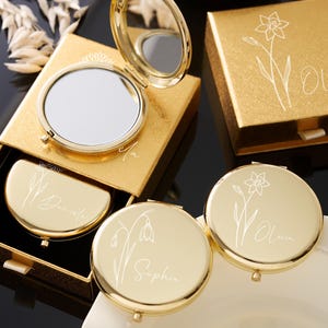 May include: Gold-toned compact mirrors and matching boxes. The mirrors are round and open to reveal a reflective surface. Each compact and box features a floral design and a name in elegant script. The boxes are square and rectangular.