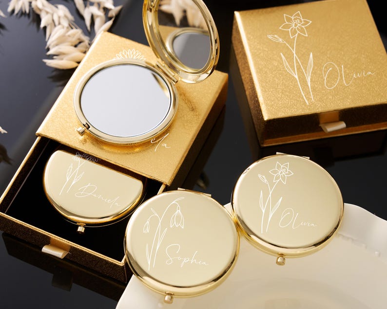 May include: Gold-toned compact mirrors and matching boxes. The mirrors are round and open to reveal a reflective surface. Each compact and box features a floral design and a name in elegant script. The boxes are square and rectangular.