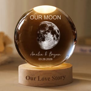 Può includere: Una sfera di vetro trasparente con un'immagine dettagliata della luna, illuminata dall'interno, poggia su una base di legno. La sfera è incisa con "OUR MOON", i nomi "Amelia & Bryan" e una data. La base recita "Our Love Story."