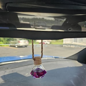 May include: A car air freshener with a clear, diamond-shaped glass bottle filled with purple liquid. The bottle is suspended by a brown and gold cord, and has a wooden cap. The air freshener hangs from the rearview mirror.