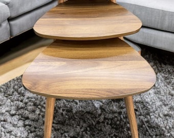Puede incluir: Un juego de tres mesas nido con tapa triangular y patas de madera. Las mesas son de madera marrón claro con veta visible. Las mesas están apiladas, la más grande en la parte inferior y la más pequeña en la parte superior. Cada mesa mide unos 60 cm de ancho.