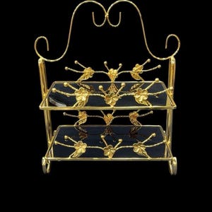 May include: A two-tiered, gold-colored serving stand with black glass shelves. The stand features ornate floral and leaf designs. The top has a decorative curved design. The stand is on a black background.
