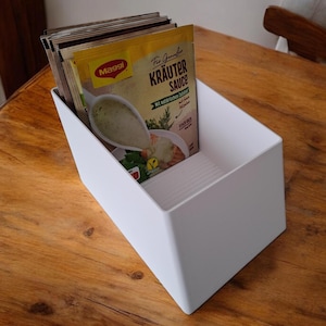 Op de afbeelding: Een witte, schuine container met verschillende pakjes Maggi Kräuter Sauce. De pakjes zijn geel en bevatten afbeeldingen van de saus. De container staat op een houten oppervlak.