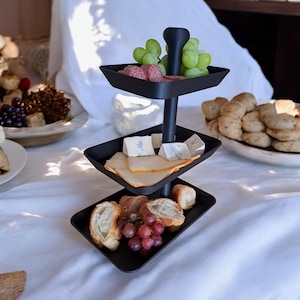 Könnte beinhalten: Ein schwarzes, dreistöckiges Serviertablett mit Essen. Die oberste Etage enthält grüne Trauben und Salami. Die mittlere Etage hat Käsescheiben und -würfel. Die untere Etage enthält Brot und rote Trauben. Das Tablett steht auf einer weißen Tischdecke.