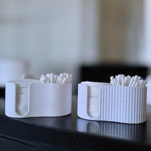 Op de afbeelding: Twee witte, minimalistische containers voor wattenstaafjes en wattenschijfjes staan op een donkere ondergrond. De ene container heeft een gladde textuur, de andere verticale ribbels. Beide zijn gevuld met witte wattenstaafjes en pads.