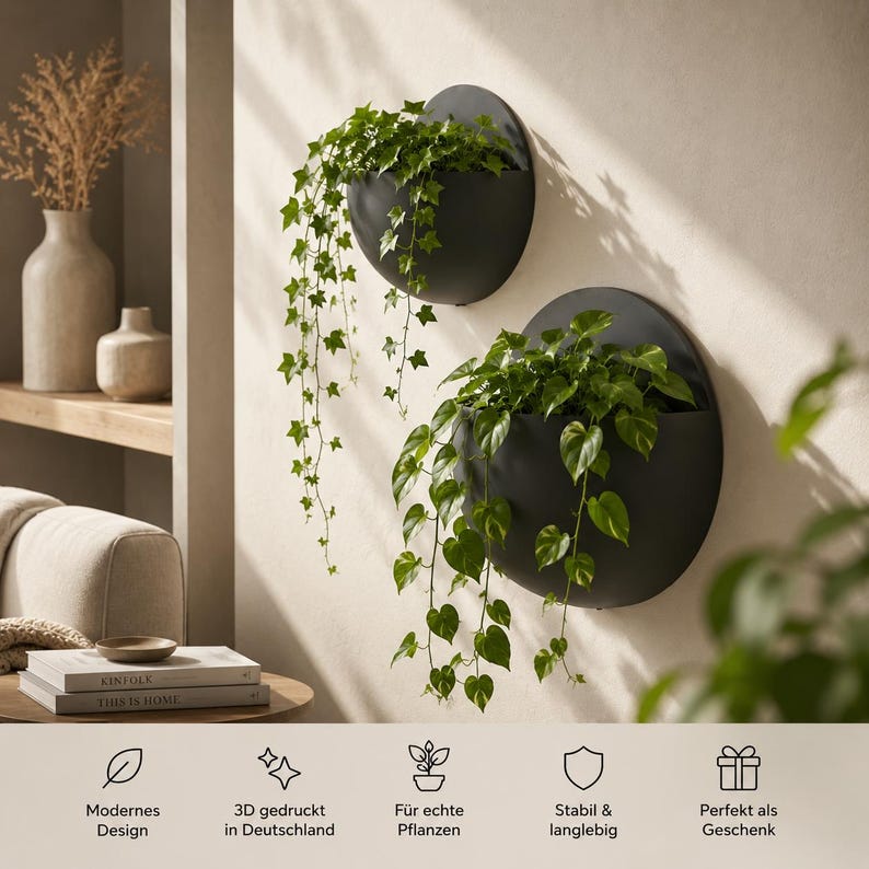 May include: Two matte black, semi-circular wall-mounted planters holding trailing green ivy and pothos plants. The planters feature a flat back and a curved front, mounted on a light-colored wall. Text at the bottom includes Modernes Design, 3D gedruckt in Deutschland, F&uuml;r echte Pflanzen, Stabil & langlebig, and Perfekt als Geschenk.