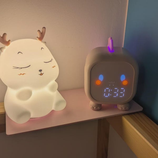 May include: A white, glowing, deer-shaped silicone night light with closed eyes and small antlers sits next to a square, light-coloured digital alarm clock featuring a purple top fin, blue and orange facial accents, and a digital display showing 0:33.