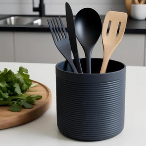 Könnte beinhalten: Ein dunkelgrauer Utensilienhalter mit geripptem Design enthält eine schwarze Gabel, ein Messer, einen Löffel und einen Holzspatel. Die Küchenutensilien sind in dem zylindrischen Behälter angeordnet, der auf einer weißen Arbeitsplatte steht. Frischer Koriander liegt auf einem Holzschneidebrett.