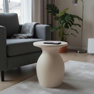 Puede incluir: Una mesa auxiliar moderna de color crema, con una parte superior redondeada y una base en forma de jarrón. Un teléfono inteligente negro descansa sobre la superficie de la mesa. La mesa está en un salón, junto a un sillón gris y una planta en maceta.