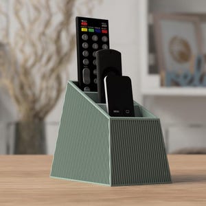 Puede incluir: Un soporte trapezoidal para mandos a distancia de color verde salvia con una textura exterior de rayas verticales. El organizador tiene tres compartimentos interiores distintos, que contienen un mando grande negro y dos mandos pequeños de streaming.