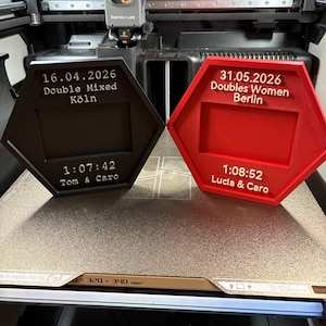 Peut inclure: Deux cadres hexagonaux imprimés en 3D en noir et rouge. Le cadre noir affiche 16.04.2026, Double Mixed, Köln, 1:07:42, et Tom & Caro. Le cadre rouge affiche 31.05.2026, Doubles Women, Berlin, 1:08:52, et Lucia & Caro.