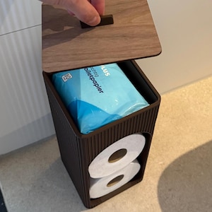 Op de afbeelding: Een donkerbruine, verticaal geribbelde opbergcontainer met een deksel in houtlook. De unit heeft een open voorvak met twee witte rollen toiletpapier en een bovenste compartiment met een blauwe verpakking met de tekst "feuchtig Toiletpapier".
