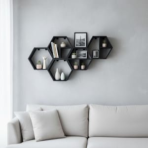 Peut inclure: Étagères murales hexagonales noires. Elles sont remplies de petites plantes en pot, de livres, de vases décoratifs et de photographies encadrées. Les étagères sont fixées sur un mur gris clair, au-dessus d'un canapé blanc.