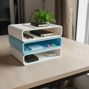 Könnte beinhalten: Ein dreistufiger Schreibtisch-Organizer in Weiß und Hellblau. Die obere und untere Ebene sind weiß, während die mittlere Ebene hellblau ist. Der Organizer enthält Notizbücher, Stifte, Haftnotizen, Büroklammern und eine tragbare Festplatte.